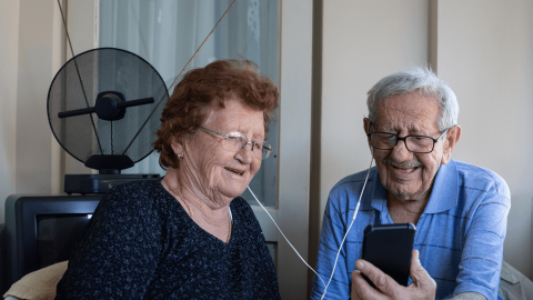 Dues persones grans comparteixen auriculars connectats a un mòbil a casa seva